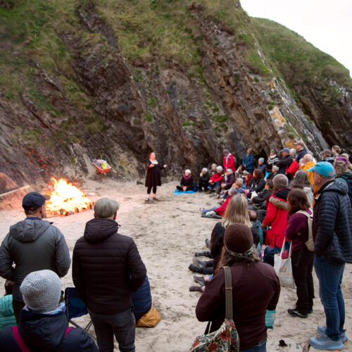 People gathered on a beach with a fire listening to someone talk